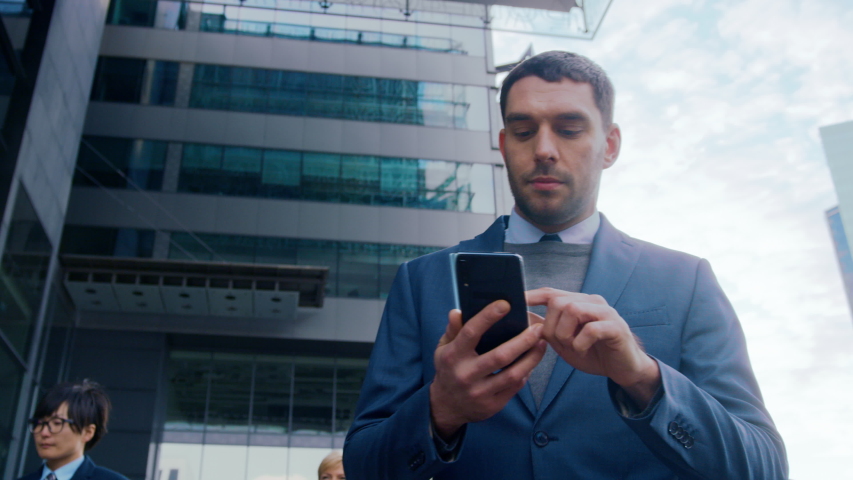 On Busy City Street Handsome Businessman Uses Smartphone with Animated Holographic Screens Show Business Graphs, Charts and Stock Market Analysis Statistics. Mock-up Mobile OS UIUX. Moving Front View Royalty-Free Stock Footage #1056263540