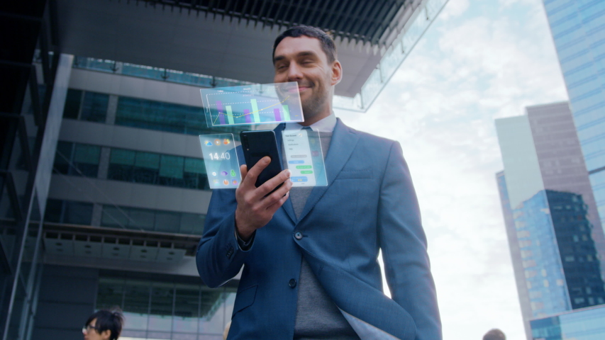 On Busy City Street Handsome Businessman Uses Smartphone with Animated Holographic Screens Show Business Graphs, Charts and Stock Market Analysis Statistics. Mock-up Mobile OS UI/UX. Moving Front View Royalty-Free Stock Footage #1056263540