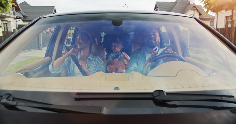 Front view of joyful beautiful parents and their excited girls smiling driving a car in the city. Young family dancing in car in summer. Concept of lifestyle, happy family, travel together.