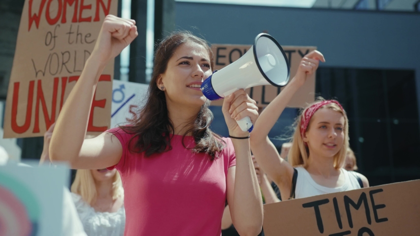 Strong woman walks in protesting crowd shouting slogans on megaphone. Group of aiming young women with placards taking part in feminist street manifestation.