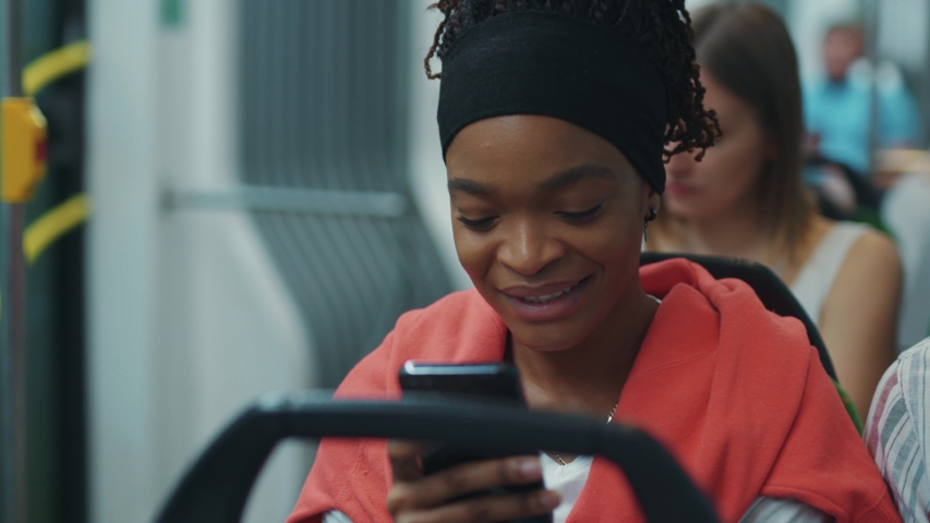 Charming afro-american woman with smartphone read messages from boyfriend smiling in the tram. Public transport. Motion. Urban people. | Shutterstock HD Video #1056264050