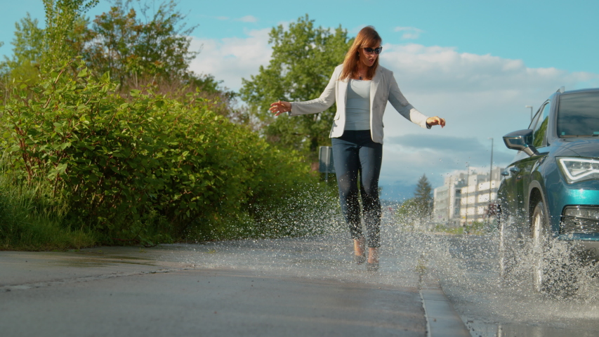 SLOW MOTION, LOW ANGLE, DOF: Unknown driver drives their blue SUV into a puddle, splashing water at young businesswoman. Unsuspecting girl walking along sidewalk gets splashed by careless car driver.