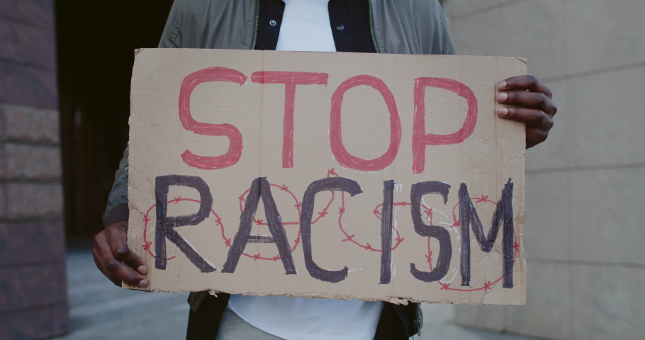 Crop view of african american person hands holding carton placard with stop racism writing. Male activist protesting at city street. Concept of human rights and racial inequality. Royalty-Free Stock Footage #1056265988