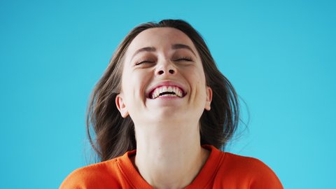 Studio portrait of smiling young woman with long hair throwing her head back into frame and laughing on blue background - shot in slow motion