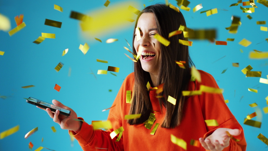 Celebrating young woman with mobile phone winning prize and showered with gold confetti in studio - shot in slow motion Royalty-Free Stock Footage #1056266165