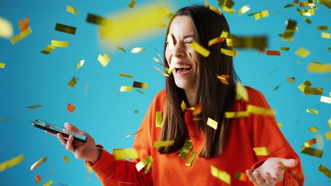Celebrating young woman with mobile phone winning prize and showered with gold confetti in studio - shot in slow motion