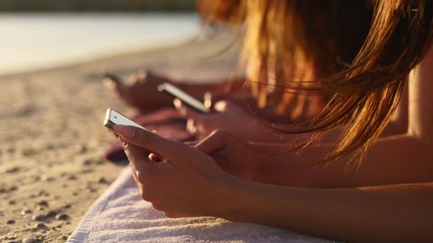 Group of millenial girls using smartphones laying together on beach towel near sea on summer sunset. Young women addicted by mobile smart phones. Always connected generation communicate via internet.