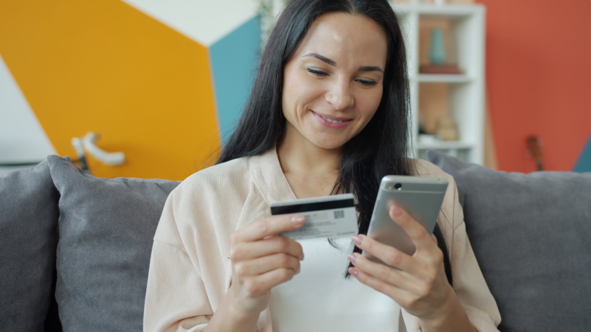 Slow motion of joyful girl shopping online holding bank card and smartphone in apartment then smiling enjoying activity. People and technology concept. Royalty-Free Stock Footage #1056267740
