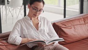 Concentrated elegant pretty asian woman in eyeglasses reading magazine while sitting on sofa at home - Powered by Shutterstock - Get 15% off with code: PIKWIZARD15