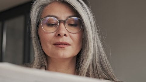 A close-up view of a beautiful mature woman wearing eyeglasses is reading a newspaper sitting at the table at home in the morning