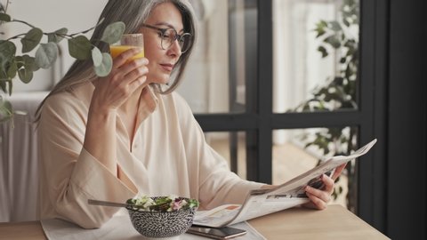 A gorgeous mature woman in eyeglasses is reading newspaper while eating breakfast sitting at the table at home
