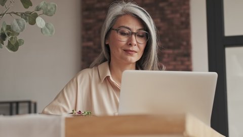 An attractive mature woman in eyeglasses is using to her silver laptop computer while eating food sitting at the table at home