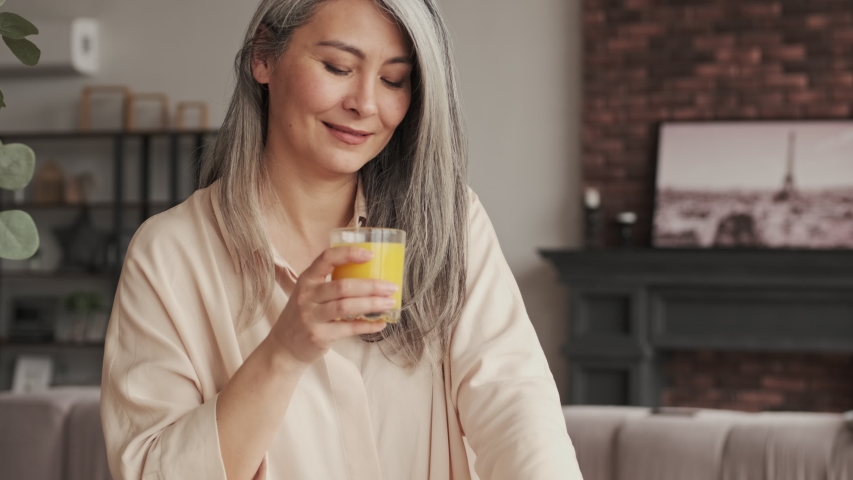 A smiling mature woman is drinking orange juice in the kitchen at home