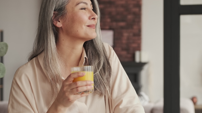 A smiling mature woman is drinking orange juice in the kitchen at home