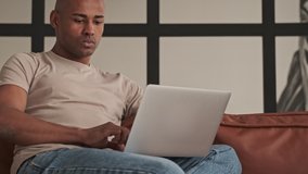 A handsome african american man is looking to his laptop sitting on the couch in living room at home