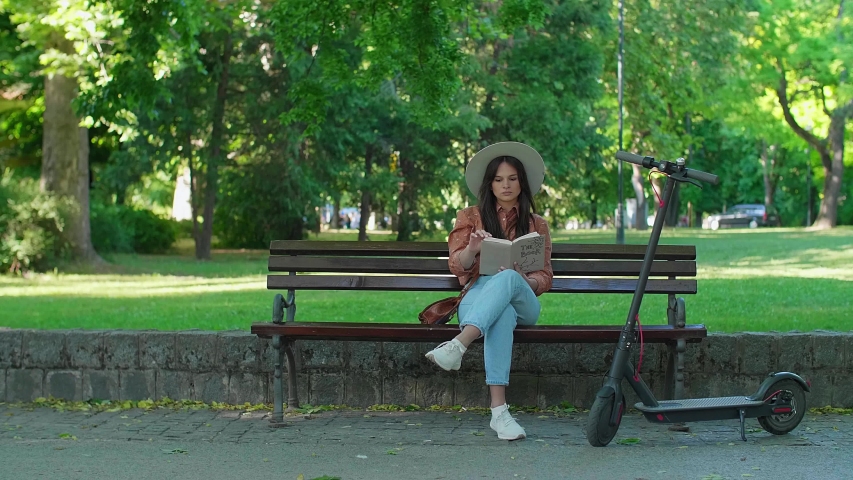 A young beautiful girl with a hat is sitting on a park bench with the book in her hands while drinking coffee. An electric scooter is parked next to her, while trees predominate in the background.