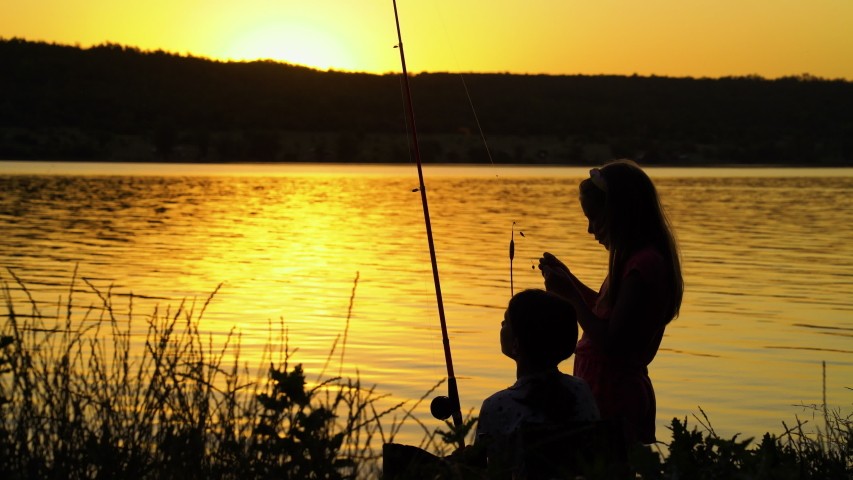 Silhouettes of children with fishing rods on river bank at sunset