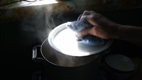Sharp sunlight to retro cooking pot with white cover and hot steam over boiling broth inside. Granny's hands open pot and put meat into hot bouillon. Hot water steam in bright light with dark shadows