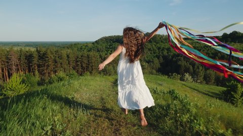 Happy young girl in a white dress and long hair running down the mountain, holding rainbow-colored ribbons fluttering in the wind