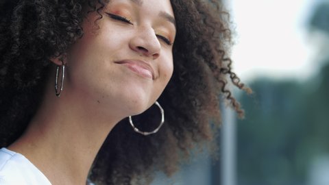 Close-up of pretty young afro american millennial woman smile with happiness joy at camera playfully winks eye with flashing gaze. Beautiful positive girl with curly hair in good mood outside in city.