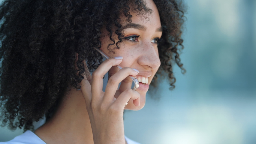 Ethnic American contented curly woman talking on phone with friends rejoicing smiling laughing white teeth, outdoors close-up. Girl student makes online order on smartphone outside, satisfied customer Royalty-Free Stock Footage #1056283940