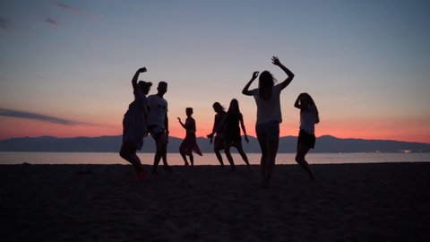 A group of friends dancing barefoot at the sea beach on amazing sunset background, crowd of happy friends having fun at the beach party, celebrating the end of summer holidays and vacation