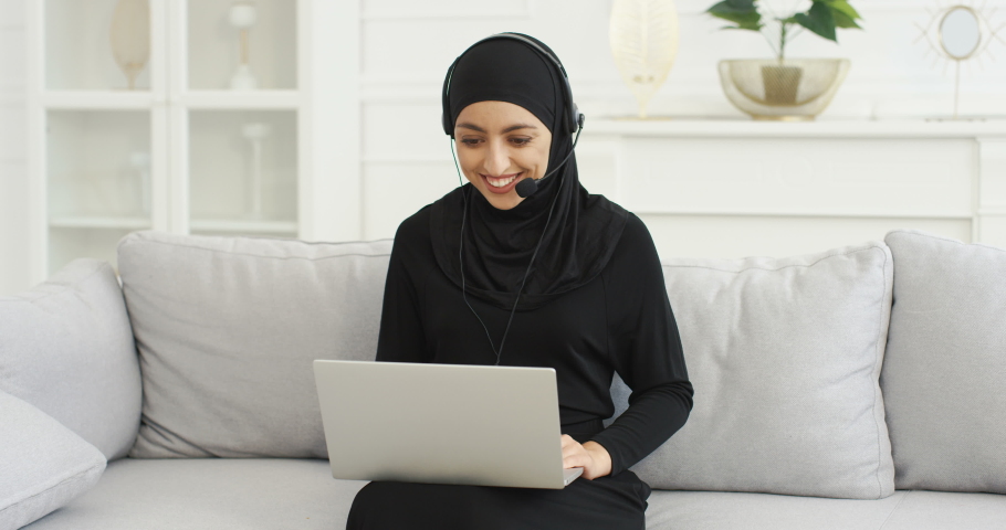 Pretty young Arabian woman blogger in headset sitting at laptop computer on couch and talking. Female vlogger in hijab working and educating. Video blogging. Home vlog studying. Muslim. Education. Royalty-Free Stock Footage #1056287765