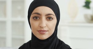 Portrait of beautiful young Arab muslim woman in black traditional hijab looking straight to camera and smiling. At home. Close up of female pretty face with smile. Arabian headscarf. Indoor.