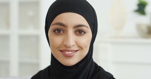 Portrait of beautiful young Arab muslim woman in black traditional hijab looking straight to camera and smiling. At home. Close up of female pretty face with smile. Arabian headscarf. Indoor.