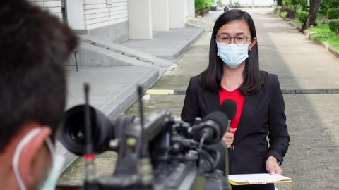 Reporter woman holding a microphone with reporting news and cameraman shooting outdoor news update while wearing  mask prevent Covid-19 or coronavirus quarantine pandemic.
