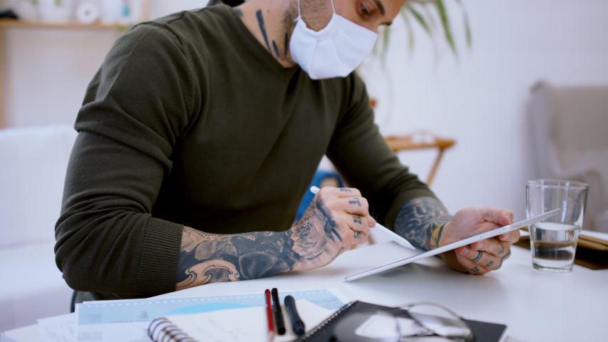 Man with face mask and tablet sitting indoors at desk in home office, working.