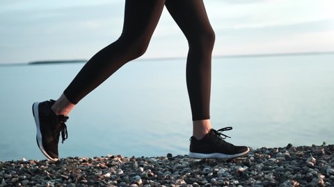 Female legs in sneakers and sportswear running on pebbles near sea at beach slow motion tracking shot. Feet of fitness woman jogging outdoor at seashore side view. Close up shot on 4k RED camera
