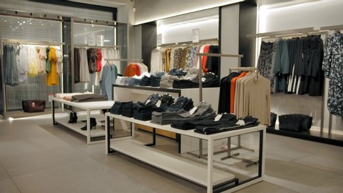 Wide vertical panning shot indoor of clothing store with modern fashionable cloth hangers and shoes. Dresses sweaters jeans, trousers tops and other clothes.