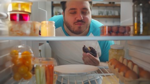 Overweight young man opening fridge and eating delicious doughnut. Obese guy with food addiction grabbing unhealthy food from refrigerator. Diet failure, unhealthy lifestyle concept