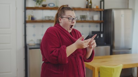 Happy fat overjoyed young woman holding mobile phone celebrating bid win standing at home. Excited overweight woman checking weight loss results in phone app
