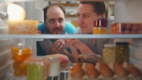 View from fridge of happy fat young couple taking fatty food from refrigerator and smiling. Stout man and woman grabbing snack from fridge. Unhealthy diet, overeating and obesity concept