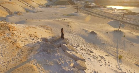 aerial view flying around girl in black dress standing at the rock on sand quarry, hot sand desert with lake