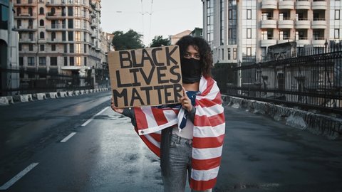 African-american protester in black bandana, wrapped in flag of USA. She showing cardboard tablet, inscription black lives matter. Standing on bridge