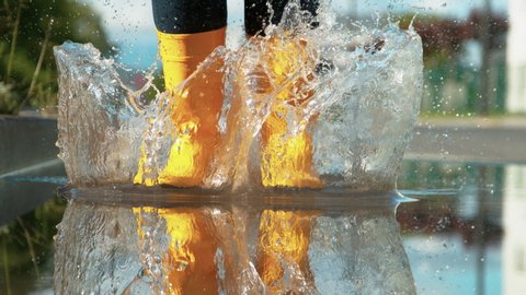 SLOW MOTION, LOW ANGLE, CLOSE UP, DOF: Unrecognizable woman in brand new yellow rubber boots jumps into the glassy puddle on the sidewalk. Carefree young woman in rain boots jumps into the big puddle.
