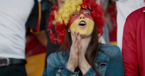 Young woman with german flag color wig and face paint clapping and chanting with group of fans for their national team. German football team supporters cheering during a match at stadium.
