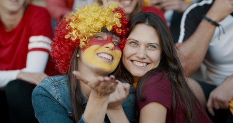 Two women sitting in fan zone with a wig and face painted in german flag colors pointing at a big screen and blowing a kiss. Germany soccer team supporters looking themselves in a big screen at stadiu