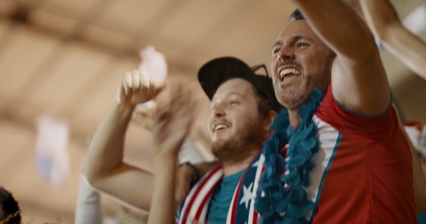 Excited fans with US flag in sports crowd celebrating victory. American soccer fans cheering over a goal in the stands.
