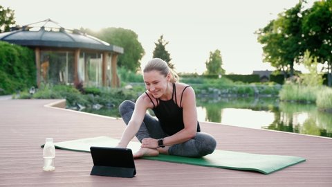 Teaching yoga online. Happy attracrive woman in sportswear looking at digital tablet and giving online yoga class while sitting on a mat in the park on a summer morning