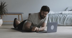 Online training. Bearded guy using stopwatch on phone, standing in plank and watching video tutorial on laptop at home