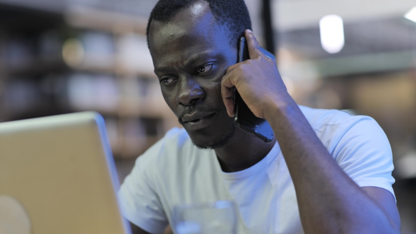 Close up of serious black young Businessman talking on the mobile phone looking at laptop in the office African freelance focus on work thinking listening on the phone in cafe | Shutterstock HD Video #1056313082