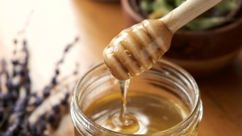 Liquid organic raw honey in glass jar with wooden honey dipper. Slow motion of golden honey flowing in a jar
