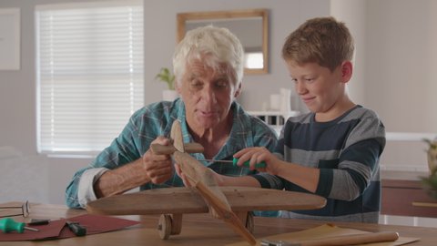Senior man helping child to screw an airplane part that they are building together. Retired old grandfather helping grandson in making wooden plane at home for school project.