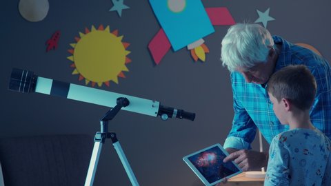 Cheerful grandfather showing stars and galaxies on digital tablet to happy grandson. Smiling old man teaching the constellations to little boy near a telescope. Child and grandpa stargazing at night.