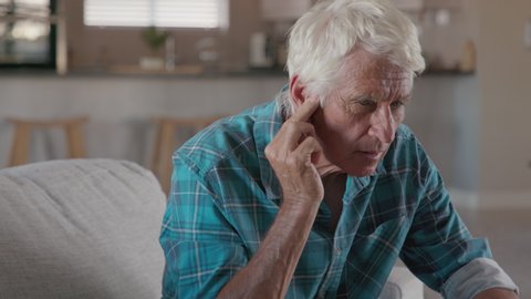 Senior man holding ears while suffering from pain. Side view of senior man with symptom of hearing loss, copy space. Mature tensed man sitting on couch with fingers near ear suffering pain.