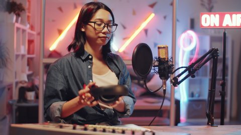 Young Asian woman putting headphones on and talking into microphone while recording podcast in home radio studio with colorful neon light and illuminated on-air sign